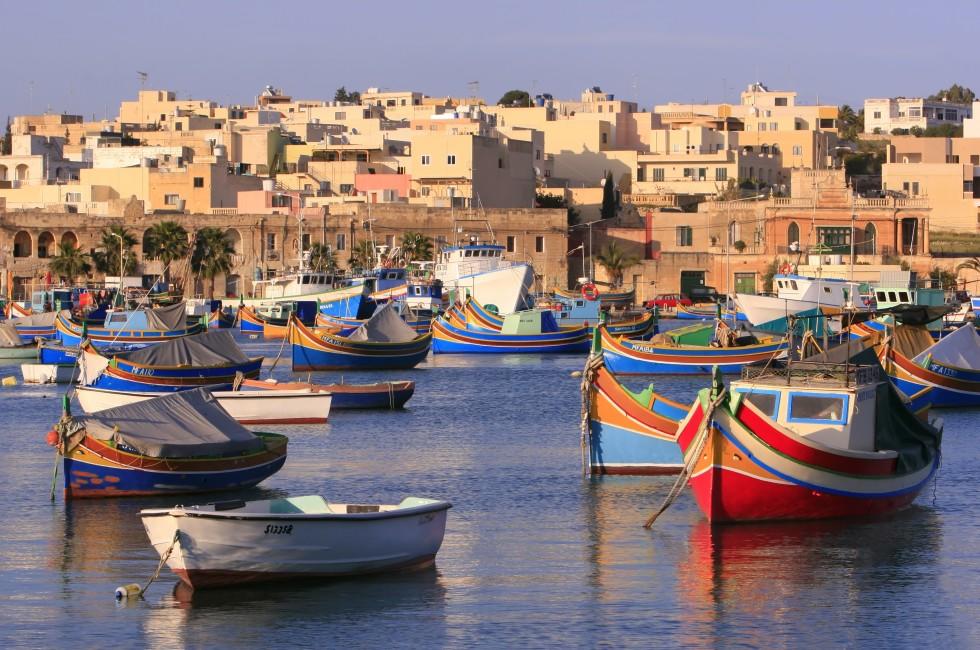 Colorful, traditional fishing boats against the backdrop of Marsaxlokk village in the mediterranean island of Malta. See portfolio for more.; Shutterstock ID 3776389; Project/Title: fodors.com destinations; Downloader: Melanie Marin